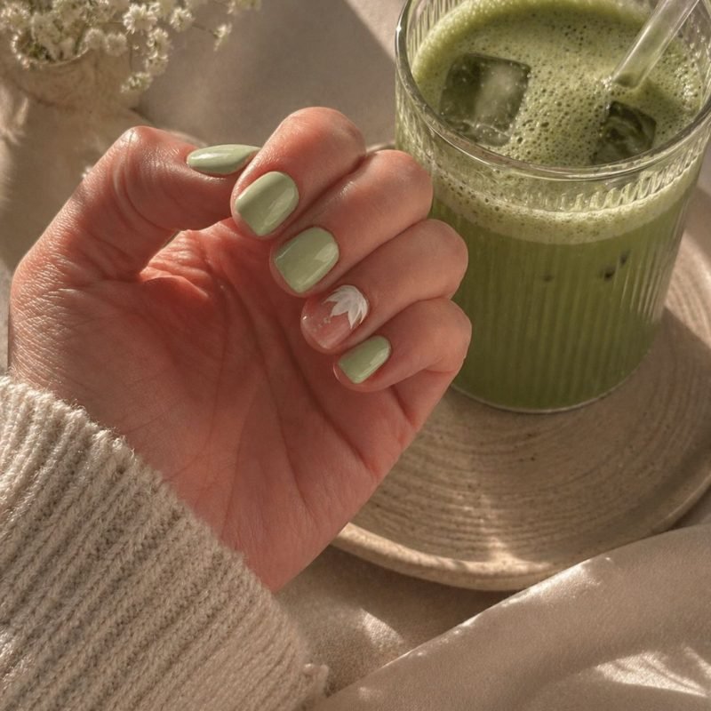 Salbeigrüne Maniküre mit weißem Blütenakzent neben Matcha-Glas